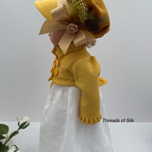 May include: A doll wearing a yellow bonnet with a large bow and feather accents, paired with a matching yellow jacket and a white eyelet dress. The outfit is reminiscent of historical fashion.