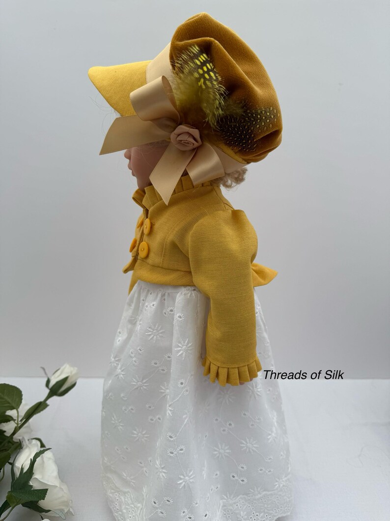 May include: A doll wearing a yellow bonnet with a large bow and feather accents, paired with a matching yellow jacket and a white eyelet dress. The outfit is reminiscent of historical fashion.