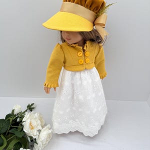 May include: A doll wearing a mustard yellow hat with a wide brim and a matching jacket with four yellow buttons. The doll is also wearing a white eyelet dress with a scalloped hem. A tan bow and sprig of greenery adorn the hat.