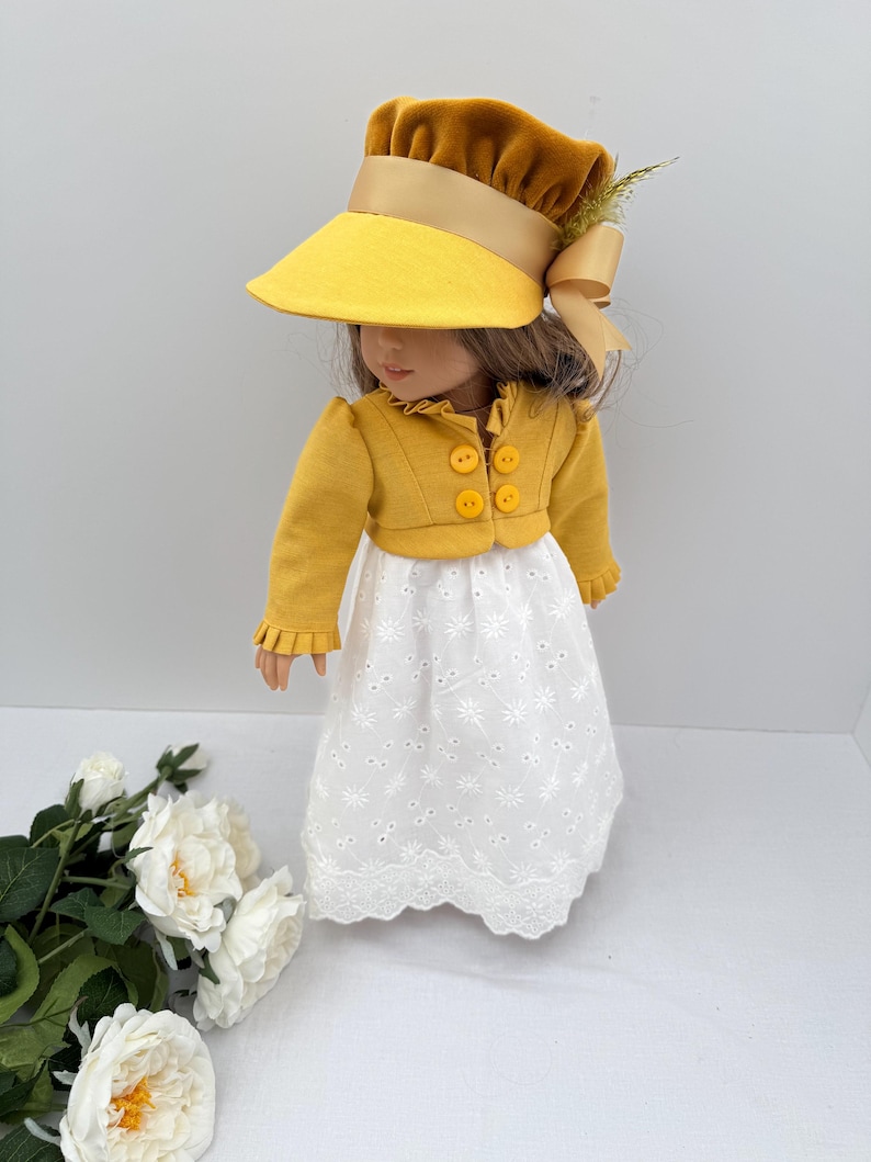 May include: A doll wearing a mustard yellow hat with a wide brim and a matching jacket with four yellow buttons. The doll is also wearing a white eyelet dress with a scalloped hem. A tan bow and sprig of greenery adorn the hat.