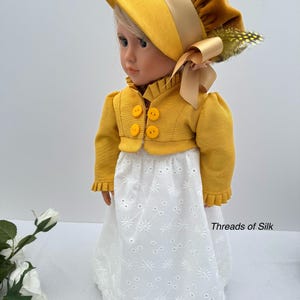 May include: A doll wearing a yellow bonnet, jacket, and white eyelet skirt. The bonnet has a wide brim and is adorned with a ribbon and feather. The jacket has three yellow buttons. The skirt has a floral pattern. The text "Threads of Silk" is visible.