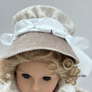 Puede incluir: Una mu&ntilde;eca con un gorro beige con una cinta blanca y detalles de perlas. El gorro tiene un patr&oacute;n texturizado y se combina con un abrigo a juego. La mu&ntilde;eca tiene el pelo rubio rizado y est&aacute; sobre un fondo claro. El texto "Threads of Silk" es visible.
