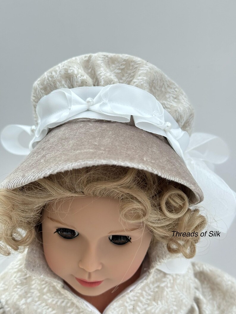 Puede incluir: Una mu&ntilde;eca con un gorro beige con una cinta blanca y detalles de perlas. El gorro tiene un patr&oacute;n texturizado y se combina con un abrigo a juego. La mu&ntilde;eca tiene el pelo rubio rizado y est&aacute; sobre un fondo claro. El texto "Threads of Silk" es visible.