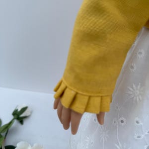 May include: A doll's arm is shown wearing a mustard yellow long-sleeved top with a ruffled cuff. The doll is also wearing a white eyelet dress with floral embroidery. The background is a soft white.