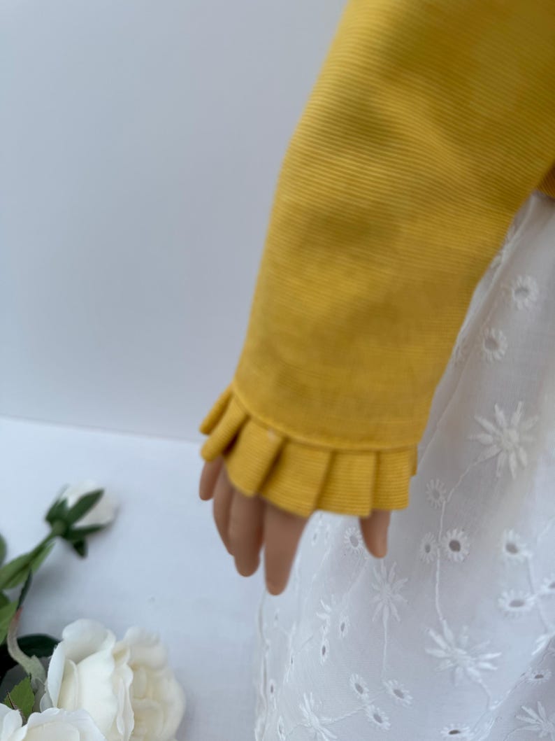 May include: A doll's arm is shown wearing a mustard yellow long-sleeved top with a ruffled cuff. The doll is also wearing a white eyelet dress with floral embroidery. The background is a soft white.