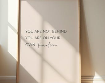 May include: A framed print with a light beige background and the quote, "YOU ARE NOT BEHIND YOU ARE ON YOUR OWN Timeline" in black text. The frame is a light brown color, and the print is leaning against a wall.