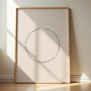 May include: A framed print with a light beige background and a wooden frame. The print features a circular text design in black that reads: "The universe responds to the frequency of your heart, not your words."