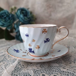 May include: A light blue teacup and saucer set with gold trim and decorative cross-shaped designs in gold and navy blue. The cup has a unique, ridged design. The set is likely for tea or coffee.