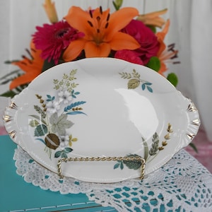 May include: White oval serving dish with a gold rim and a floral design featuring green leaves, white flowers, and brown accents. The dish is on a white crocheted doily.
