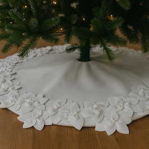 May include: A white Christmas tree skirt with a floral design. The skirt is made of a soft fabric and features white fabric flowers with pearl accents around the edge. The skirt is under a green Christmas tree.