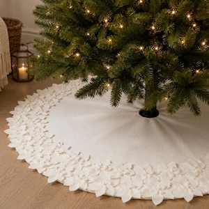 May include: A circular white Christmas tree skirt featuring a decorative border of layered, petal-like fabric cutouts. The skirt is positioned at the base of a green pine tree adorned with small, warm-toned glowing string lights.