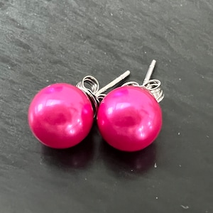 Può includere: Un paio di orecchini a bottone rotondi rosa acceso. Gli orecchini hanno una finitura lucida, simile alla perla, e sono montati su perni d'argento. Gli orecchini sono esposti su una superficie scura e strutturata.