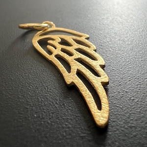 Könnte beinhalten: Ein goldfarbener Anhänger aus Metall in Form eines Engelsflügels. Der Flügel hat ein detailliertes, filigranes Design und eine gebürstete Oberfläche.
