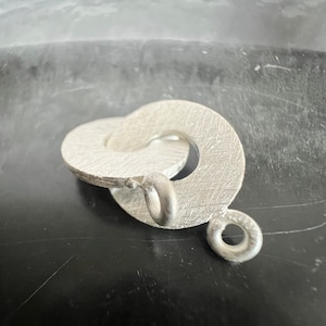 Könnte beinhalten: Ein silberner Anhänger mit einem Doppel-Loop-Design und einem kleinen Ring. Der Anhänger hat eine gebürstete Oberfläche.