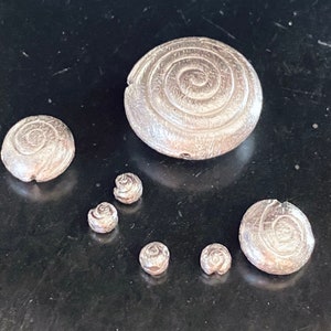 Könnte beinhalten: Ein Satz von sieben silberfarbenen Metallperlen mit einem Spiralmuster. Die Perlen sind in verschiedenen Größen und auf einer schwarzen Oberfläche angeordnet.