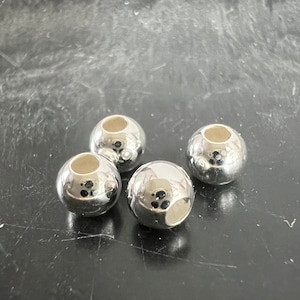 Pode incluir: Quatro contas de metal prateadas com um furo no centro. As contas são redondas e têm um acabamento brilhante.