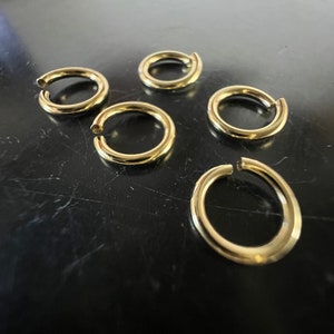 Op de afbeelding: Zes goudkleurige metalen sprongringen, elk met een ronde vorm, zijn gerangschikt op een zwart oppervlak.