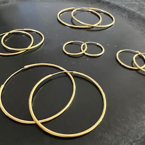 Könnte beinhalten: Eine Sammlung goldfarbener Creolen in verschiedenen Größen. Die Ohrringe sind auf einer dunklen Oberfläche angeordnet, wodurch ihr kreisförmiges Design hervorgehoben wird. Das Bild zeigt die unterschiedlichen Durchmesser der Creolen, von klein bis groß.
