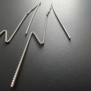 Może przedstawiać: Para srebrnych kolczyków typu threader. Każdy kolczyk ma delikatny, skręcony łańcuszek, który przechodzi w prosty, smukły słupek. Kolczyki są prezentowane na ciemnym tle, podkreślając ich metaliczny połysk.