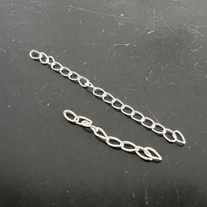 May include: Two silver chains, one longer than the other, are laid out on a dark gray surface.