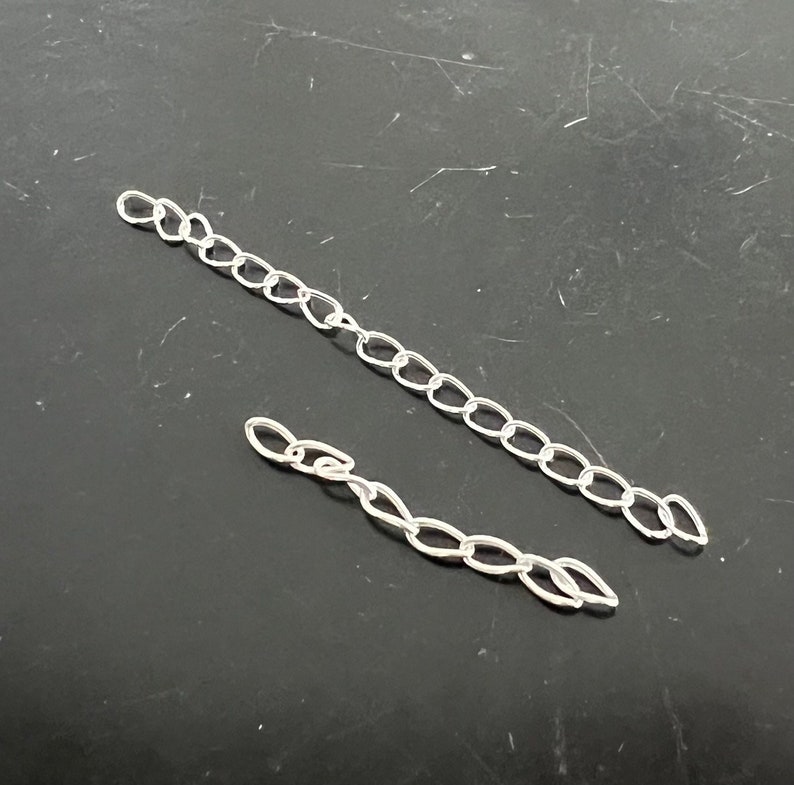 May include: Two silver chains, one longer than the other, are laid out on a dark gray surface.