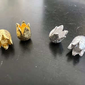 Pode incluir: Quatro pequenas formas de botão de flor de metal decorativo. Duas são douradas e duas são prateadas. Os botões de flor estão abertos e têm uma superfície texturizada.