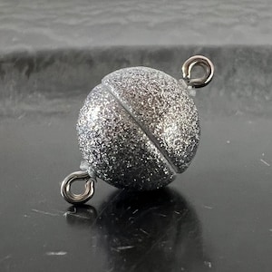Op de afbeelding: Zilveren magneetsluiting met een glinsterende afwerking. De sluiting bestaat uit twee ronde helften die met een magneetsluiting aan elkaar worden bevestigd.