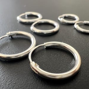 Peut inclure: Plusieurs boucles d'oreilles créoles argentées de différentes tailles sont disposées sur une surface sombre. Les boucles d'oreilles ont une finition brillante et métallique et un design simple et classique. Les créoles sont rondes et semblent être en métal massif.