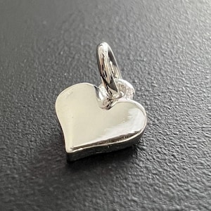 Könnte beinhalten: Ein kleiner, silberner herzförmiger Anhänger mit einer Schlaufe für eine Kette. Der Anhänger ist glatt und reflektierend, vor einem dunklen Hintergrund. Die Herzform ist klar definiert, was auf ein Schmuckstück hindeutet.