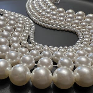 Könnte beinhalten: Eine Sammlung weißer Perlen in verschiedenen Größen, die in einem Kreis angeordnet sind. Die Perlen sind glatt und glänzend und erzeugen einen luxuriösen und eleganten Look.