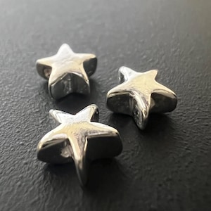 Może przedstawiać: Trzy srebrne zawieszki w kształcie gwiazd ułożone na ciemnej powierzchni. Każda gwiazda ma zaokrąglony, trójwymiarowy kształt z pięcioma ramionami. Zawieszki wydają się być wykonane z metalu i mają błyszczącą, odblaskową powierzchnię.