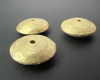 May include: Three gold-colored, textured, disc-shaped beads with a small hole in the center. The beads have a rough, organic texture and are arranged on a dark surface. The beads appear to be made of metal.