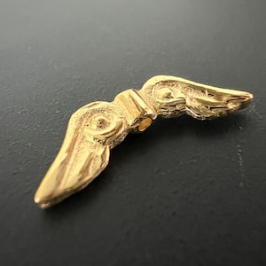 Może przedstawiać: Złoty metalowy wisiorek w kształcie skrzydła z misternymi detalami. Skrzydło jest zakrzywione i ma fakturowaną powierzchnię.