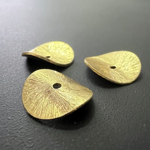 Może przedstawiać: Trzy złote metalowe elementy dekoracyjne z fakturowaną powierzchnią i otworem w środku. Elementy mają kształt łzy.