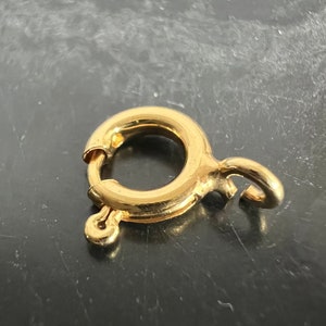 Könnte beinhalten: Vergoldeter Metallverschluss mit einem runden Ring und einem Haken. Der Verschluss ist ein gängiges Schmuckelement, das für Halsketten, Armbänder und anderen Schmuck verwendet wird.