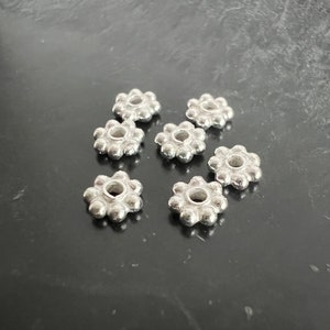 Pode incluir: Sete contas de metal prateadas em forma de flor com um furo no centro. As contas estão dispostas em semicírculo sobre uma superfície cinza escura.