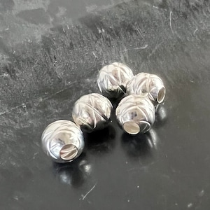 Op de afbeelding: Zes zilveren metalen kralen met een getextureerd oppervlak. De kralen hebben een gat in het midden om aan te rijgen.