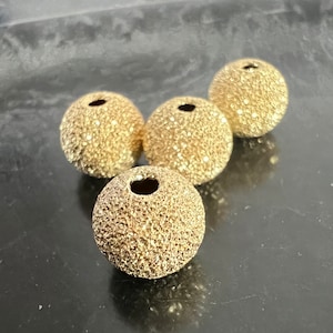 Może przedstawiać: Cztery okrągłe koraliki w kolorze złotym o fakturowanej powierzchni. Koraliki mają otwór w środku.