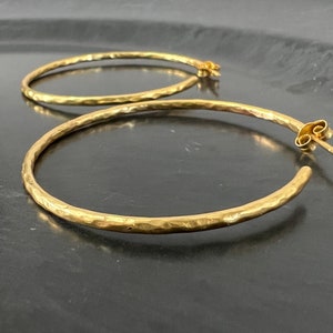 Peut inclure: Une paire de boucles d'oreilles créoles en or avec une texture martelée. Les boucles d'oreilles sont grandes et ont un design simple et élégant.