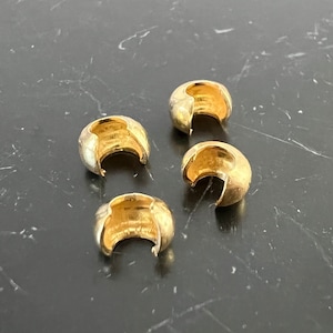 Könnte beinhalten: Vier goldfarbene Metallperlenkappen mit einer glatten, abgerundeten Oberfläche und einem leicht geöffneten, geschwungenen Design. Die Kappen sind auf einer dunkelgrauen Oberfläche angeordnet.