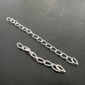 May include: Two silver chains, one longer than the other, are laid out on a dark gray surface. The chains are made of oval links.