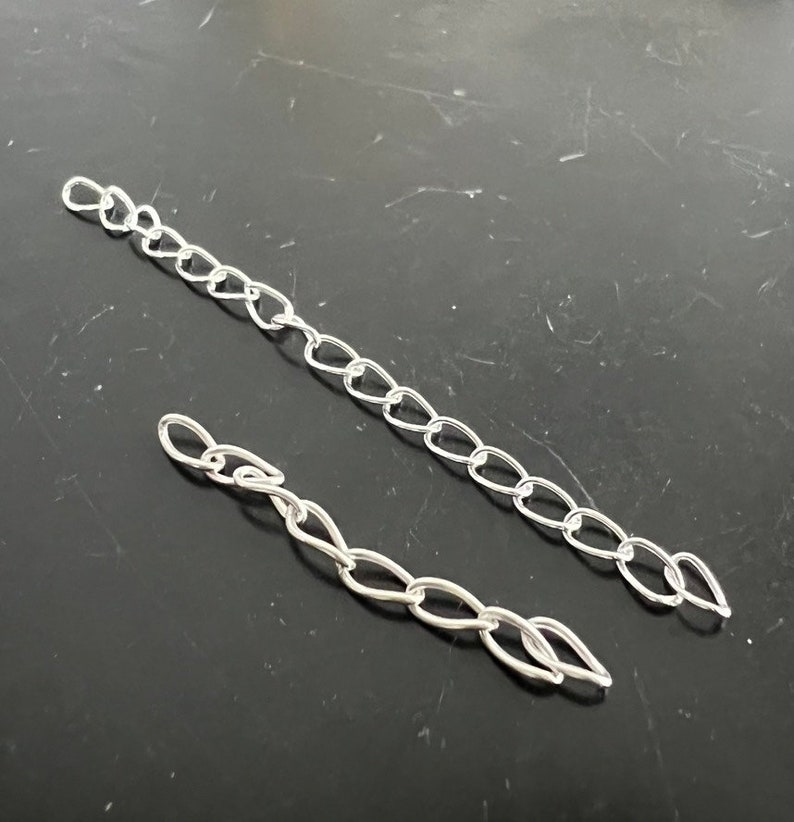 May include: Two silver chains, one longer than the other, are laid out on a dark gray surface. The chains are made of oval links.