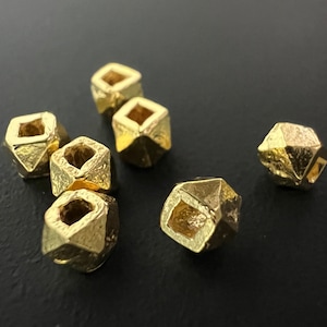 Może przedstawiać: Siedem złotych metalowych koralików o kwadratowym kształcie z otworem w środku. Korale mają fakturowaną powierzchnię i są rozrzucone na czarnym tle.