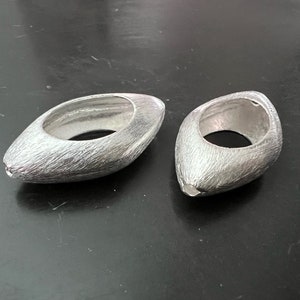 Könnte beinhalten: Zwei silberne, ovale Ringe mit einer strukturierten Oberfläche. Die Ringe sind offen und haben einen glatten, runden Innenraum.
