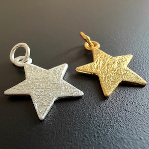 Könnte beinhalten: Zwei sternförmige Anhänger, einer silberfarben und einer goldfarben, mit strukturierten Oberflächen. Der silberne Anhänger hat oben eine kleine Schlaufe, während der goldene Anhänger eine kleine Schlaufe und einen Verbindungsring hat. Beide liegen auf dunklem Untergrund.