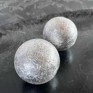 Op de afbeelding: Twee zilveren metalen bollen met een getextureerd oppervlak.
