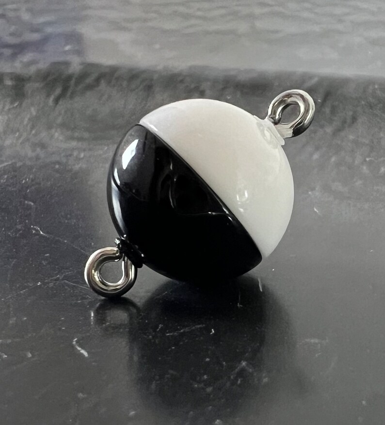 May include: A black and white glass sphere with a silver metal loop on each side. The sphere is half black and half white.