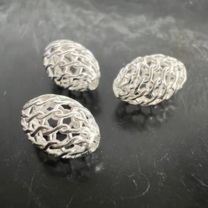Könnte beinhalten: Drei silberne, durchbrochene, ovale Perlen. Die Perlen bestehen aus einem filigranen, miteinander verwobenen Drahtmuster.