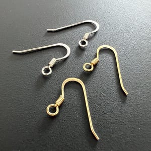 Könnte beinhalten: Nahaufnahme von vier Ohrringhaken. Zwei sind silberfarben und zwei goldfarben. Jeder Haken hat ein gebogenes Design mit einer Spirale und einer kleinen Schlaufe am unteren Ende. Die Haken befinden sich auf einer dunkelgrauen Oberfläche.