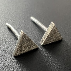 Peut inclure: Une paire de boucles d'oreilles clous argentées. Chaque boucle d'oreille présente un design triangulaire texturé fixé à une tige droite. Les boucles d'oreilles sont présentées sur une surface sombre.
